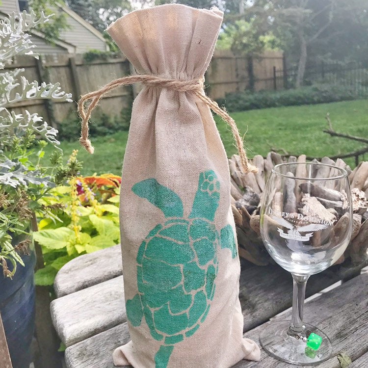 Brown burlap bag with a hand stenciled aqua blue sea turtle standing up with a brown cord around the top, wine glass to the right of it on a a picnic table, yard in the background. 