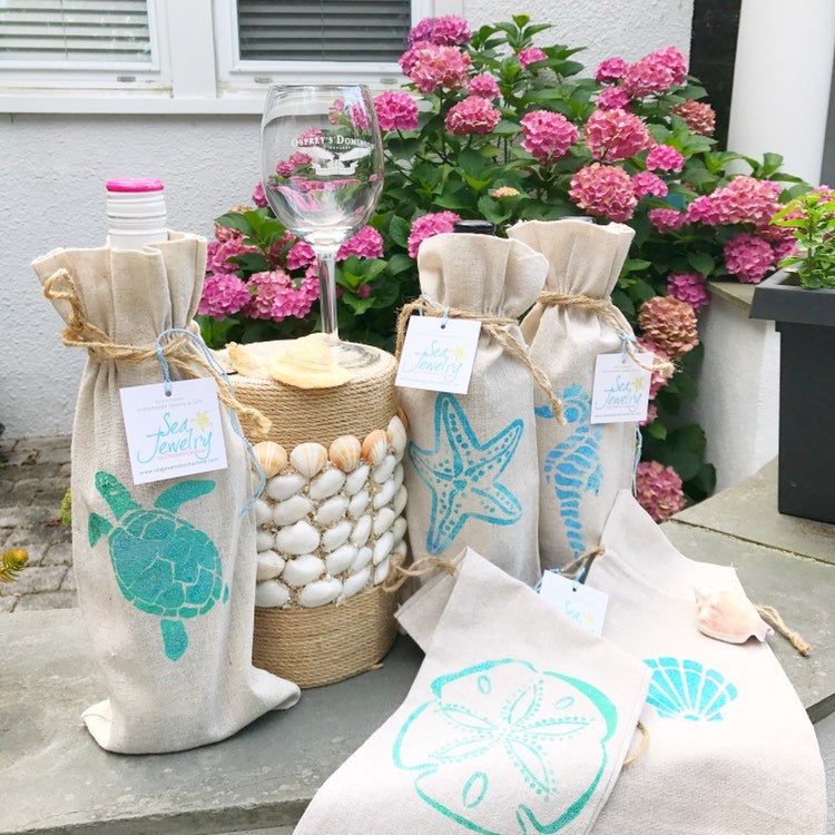 Three brown gift bags, hand stenciled, one on the right with a sea turtle hand stenciled, a wine glass displayed to the right, and a bag with hand stenciled starfish, another next to it with a hand stenciled seahorse. A pot of pink flowers in background, white building behind. Two brown bags lying empty on table. seadollar stencile. All stencils are aqua blue .