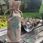 Load image into Gallery viewer, Brown burlap bag with a hand stenciled sea horse standing up with a brown cord around the top, wine glass to the right of it on a a picnic table, yard in the background. 

