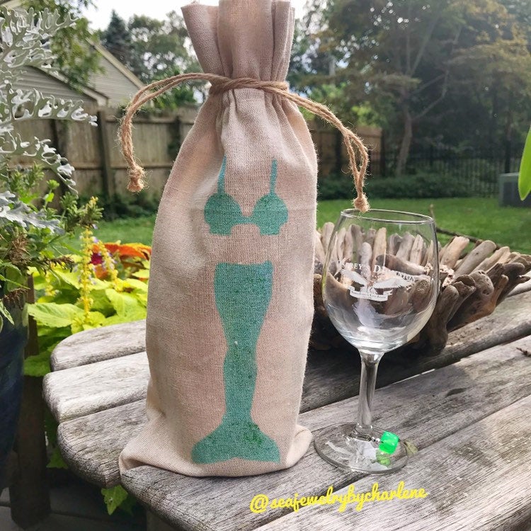 Brown burlap bag with a hand stenciled sea horse standing up with a brown cord around the top, wine glass to the right of it on a a picnic table, yard in the background. 