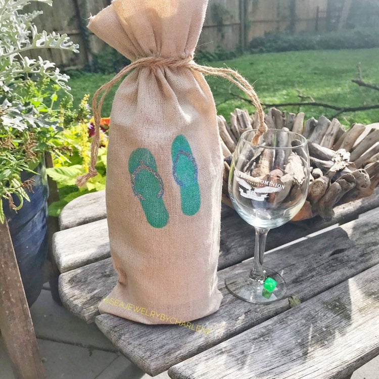Brown burlap bag with a hand stenciled aqua blue flip flops,  standing up with a brown cord around the top, wine glass to the right of it on a a picnic table, yard in the background. 